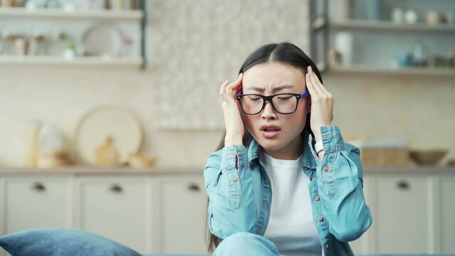 Close-up of young asian woman feeling headache while sitting on sofa in living room Tired female wearing glasses and casual clothes is giving herself a head and bridge of nose massage with closed eyes