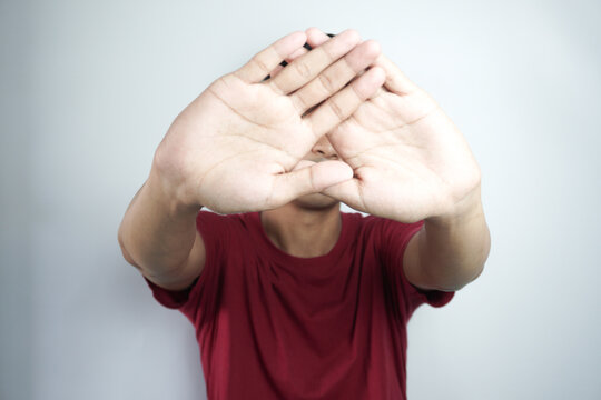 young asian man putting hand up front to stop camera, refusing photos or pictures against flat color wall