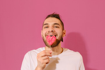 Young Caucasian man licks sweet red heart-shaped lollipop with tongue and enjoys it. Isolated on...