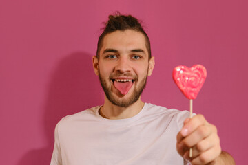 Young Caucasian man with beard shows tongue and teases sweet red lollipop in shape of heart. Isolated on pink background. Full-face portrait. Concept of Valentine's Day.