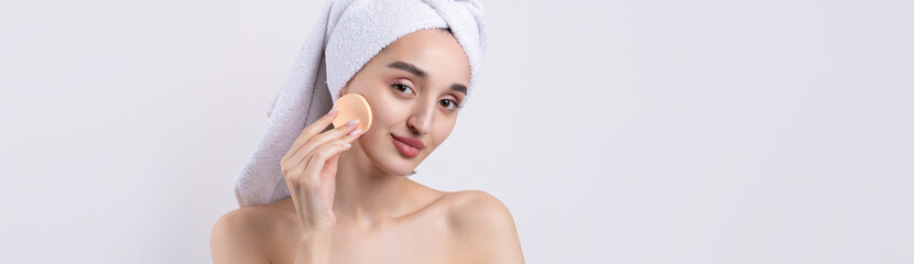 Beautiful girl with thick eyebrows and perfect skin at white background, towel on head, beauty photo. Holds a cosmetic sponge.