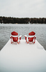 Red adirondack muskoka chairs on end of snowy dock in winter.