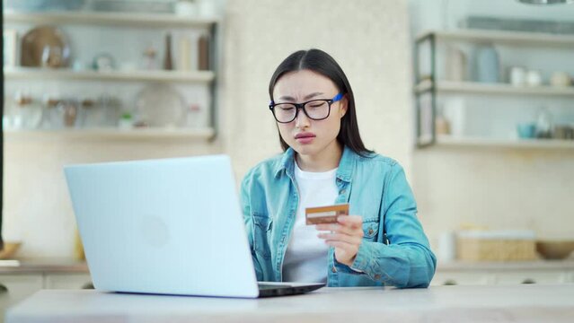 Sad asian woman unsuccessfully typing credit card number on laptop while sitting at desk at home. Young female in glasses is unhappy and upset because she made a mistake while working on the computer