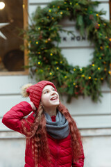 Young happy cheerful smiling girl in red jacket and knitted hat posing with hand on her head outdoors, looking up and laughing, big fir-tree cristmas wreath with lights on building wall background