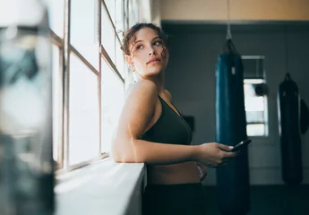 Fototapete Rund Thinking, phone and fitness with a sports woman by a window, standing in the gym during an exercise workout. Health, idea and a female athlete using social media or an app to track her training © K.A./peopleimages.com