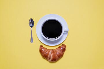 Overhead view of black coffee, spoon and croissant on yellow table