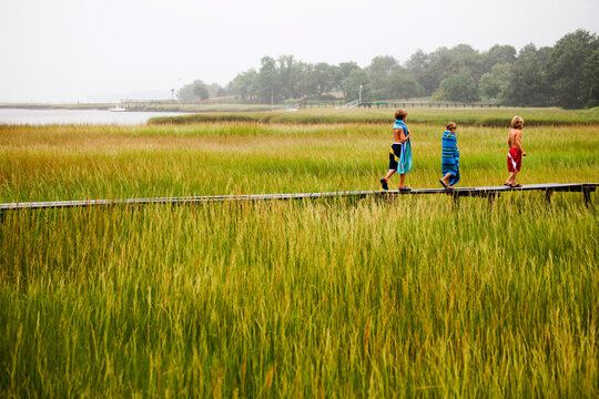 Three young children walk on a raised boardwalk over lush marsh grasses after swimming in a small inlet.