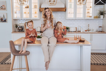 Pregnant Swedish mom sitting on a kitchen table with little daughters dressed in beige casual clothes. Daughters drinking milk. Maternity and pregnancy. Family, domestic leisure. Parenthood.