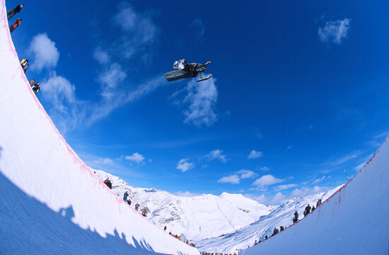 Snowmobile daredevil jumps over snowboard halfpipe.