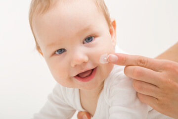 Young adult mother finger applying white moisturizing cream on baby boy cheek on light gray background. Daily care about face skin. Closeup. Front view. Happy smiling infant. - obrazy, fototapety, plakaty