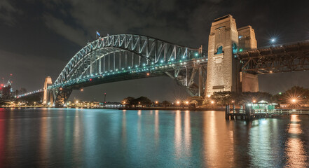 Fototapeta na wymiar Sydney Harbour Bridge at Night. Long Exposure. Cityscape In Background. Flowing Sky and Reflection on Water. Australia