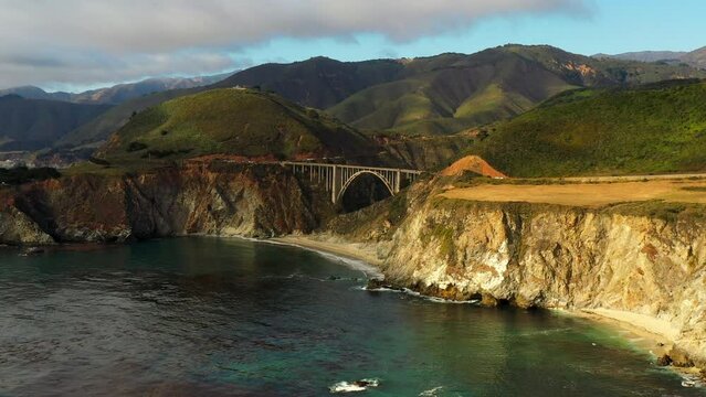 Aerial Panning Shot Of Bixby Creek Bridge Amidst Rock Formations, Drone Flying Over Sea Under Clouds - San Francisco, California