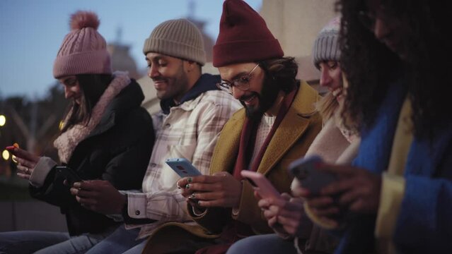 Group of young mixed race friends with mobile phones. Technology addicted people using phone at night outdoors. Concept of social media connection, friendly, app, hipster, millennial. Winter time.