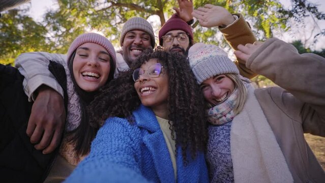 Winter time smiling selfie of a happy group of multicultural friends in video call with phone looking at camera. Cheerful multi-ethnic young diverse people having fun together. Colleague hanging out.