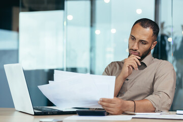 Pensive serious businessman reading financial report, hispanic businessman holding document in hands looking disappointed, working inside modern office with laptop behind paper work. - Powered by Adobe