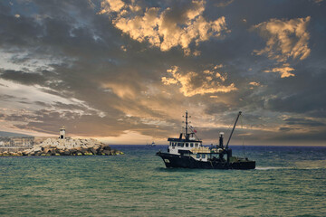 Fototapeta na wymiar View of a fishing boat returning from the sea to land and a view of the small harbor, lighthouse and gorgeous cloudy dramatic sky.