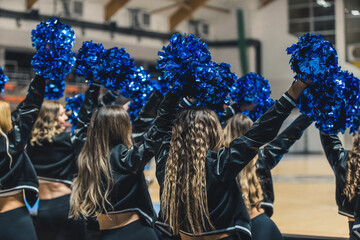 A group of cheerleaders are captured in mid-performance, their blue pom-poms waving in the air as they cheer on their team during a match. The young women are full of energy and excitement. - obrazy, fototapety, plakaty