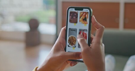 Person browsing restaurant menu online from home. Close up female hand scrolling food delivery app seated at sofa in living room. Comfortable person ordering from smartphone