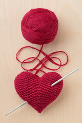 Crocheted amigurumi pink red heart with a crocet hook on a wooden background with a skein of yarn. Valentine's day