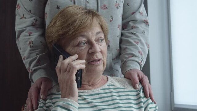 An elderly woman is talking on the phone with tears in her eyes. Daughter supports her by hugging her shoulders. Family values and friendly support