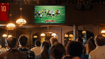 Group of American Football Fans Watching a Live Match Broadcast in a Sports Pub on TV. People...