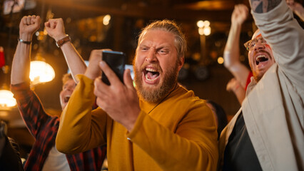 Excited Masculine Man Holding a Smartphone, Feeling Nervous About the Sports Bet He Put on a...
