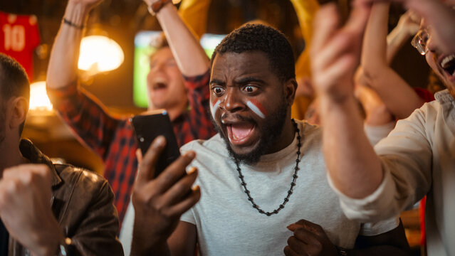 Portrait of an Excited Young Black Man Holding a Smartphone, Anxious About a Sports Bet on His Favorite Soccer Team. Lively Successful Emotions When Football Team Scores a Winning Goal.