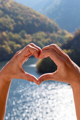 Heart shape, made by hands of women, on background of beautiful nature landscape - blue water surface of mountain river and forested green mountains. Love for nature, travel, earth day concept