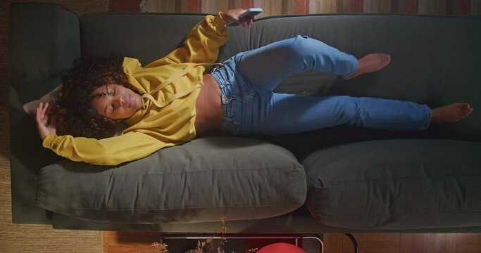 A black woman lying on sofa and checking smartphone device. An African American adult girl wearing yellow blouse staring at phone screen at night. Person relaxing