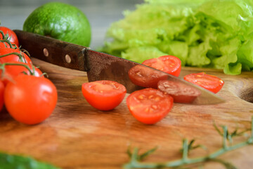 Closeup kitchen knife, sliced tomatoes on wooden cutting board on background of green vegetables indoors. Food cooking background. Ingredients for preparation salad dishes. Healthy food concept
