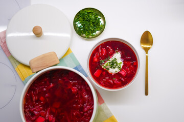 A pot of red Ukrainian borscht on a light background with a portion of soup in a bowl with sour cream and greens with a golden spoon