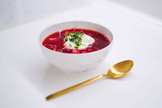 White bowl of red soup with sour cream. Red traditional borscht on a light background with a golden spoon