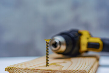 Metal self-tapping screw is screwed into the wooden board against the background of an electric screwdriver.