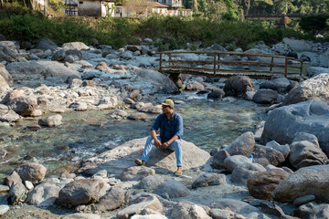 A male solo traveler sitting on a rock beside river enjoying the beauty of nature.