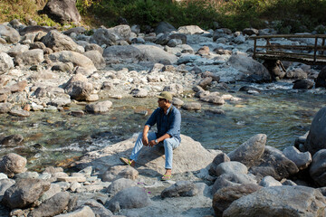A male solo traveler sitting on a rock beside river enjoying the beauty of nature.