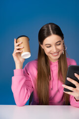 Positive teen girl holding coffee to go and using smartphone isolated on blue.