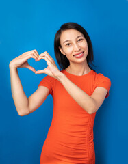 In love. Romantic symbol smiling beautiful slim Chinese Asian woman looking at camera and smile. Heart shape with hands.  Vivid orange dress and blue background. St Valentine symbol