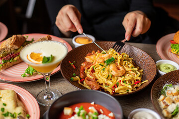 Human with fork and knife eat pasta with shrimps from plate on table full of delicious food. Hot dishes and alcoholic drink on festive dinner. Nourishing food, restaurant, beautiful serving food