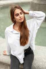 Beautiful fashionable young woman model in stylish casual clothes with a white shirt and black jeans sit and rest near the fountain in the city