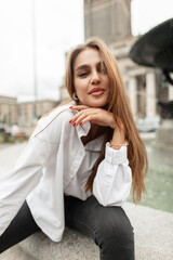 Happy stylish pretty woman with smile in casual elegant fashion outfit with white shirt sits and poses in the city