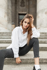 Stylish beautiful young girl vogue model in elegant streetwear with a white shirt and black jeans sits on the steps in the city near vintage buildings with columns