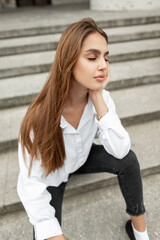 Stylish beautiful young woman model in fashionable business clothes with a shirt sits and relaxes on the steps of the city