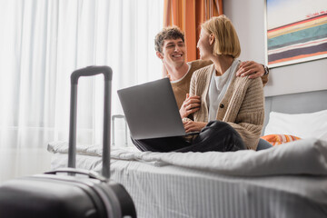 happy young couple looking at each other while sitting with laptop on bed in hotel room.