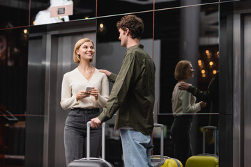 young man touching shoulder of pleased girlfriend with smartphone near suitcases in hotel lobby.
