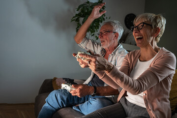 Senior couple sitting on sofa and playing video game on console. They're holding a game pad and challenge each other to win.