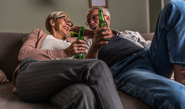 Senior couple sitting on sofa and drinks beer. They're having pleasant conversation and joying in an evening tv show.
