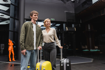 Fototapeta na wymiar young man with smartphone and happy blonde woman looking away near travel bags in lobby of modern hotel.