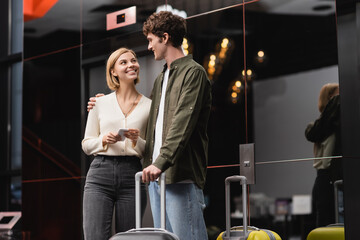 blonde woman with cellphone smiling at young boyfriend near luggage in modern hotel.
