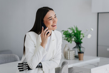 Cheerful gorgeous brunette girl with disability talking by phone, smiling looking away, laughs. Toothy smiling young Italian woman uses bionic hand, laughing. Mockup, cyber arm. Orthopaedics