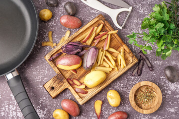Wooden board with cut and whole raw potatoes, frying pan, peeler and herbs on grunge background